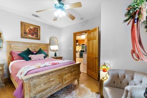 Une chambre avec un lit en bois, un ventilateur de plafond, une table de chevet avec une lampe, une chaise et une porte donnant sur la cuisine.