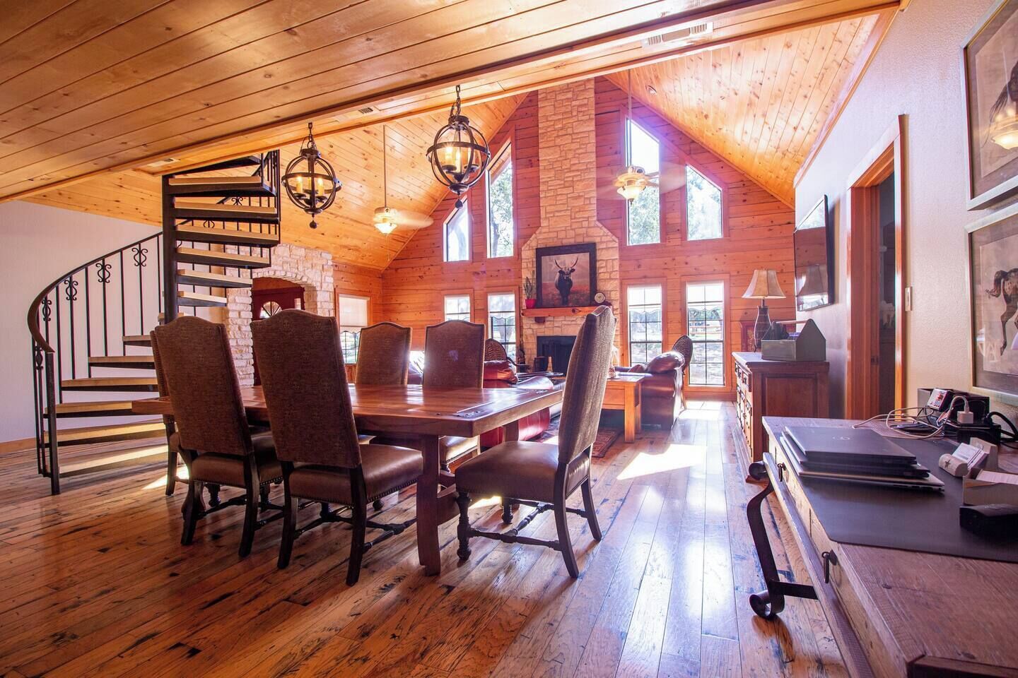 A spacious loft-style room with a high ceiling, wooden floors, a dining area with a long table and chairs, a fireplace, and large windows allowing natural light.