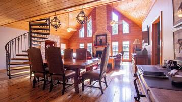 A spacious loft-style room with a high ceiling, wooden floors, a dining area with a long table and chairs, a fireplace, and large windows allowing natural light.