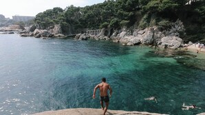 A man standing on a rocky outcrop overlooking a clear blue sea with swimmers in the water. - Lloret de Mar Camping (Lloret de Mar)
