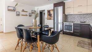 Eine moderne Küche mit Edelstahlgeräten, ein Essbereich mit Glastisch und schwarzen Stühlen sowie ein Wohnbereich mit großem Fenster.