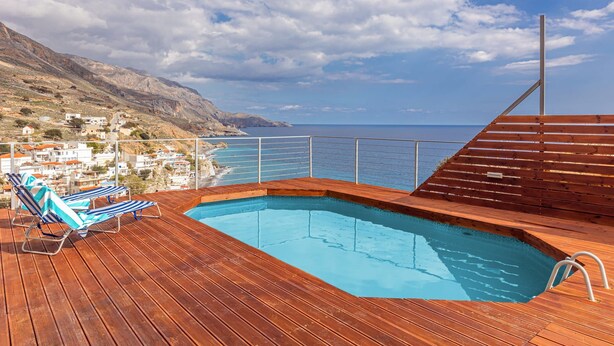 Eine Holzterrasse mit Swimmingpool, Liegestühlen und Blick auf das Meer und die Hügel.