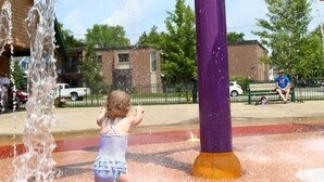 A water play area with a child playing, featuring a water fountain structure with multiple colored nozzles.