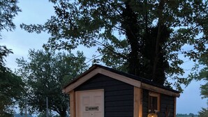 A small wooden outhouse with a white door, a small window, and a porch with four posts.