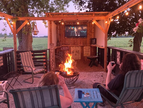 An outdoor seating area with a fire pit, wooden furniture, and a TV screen.