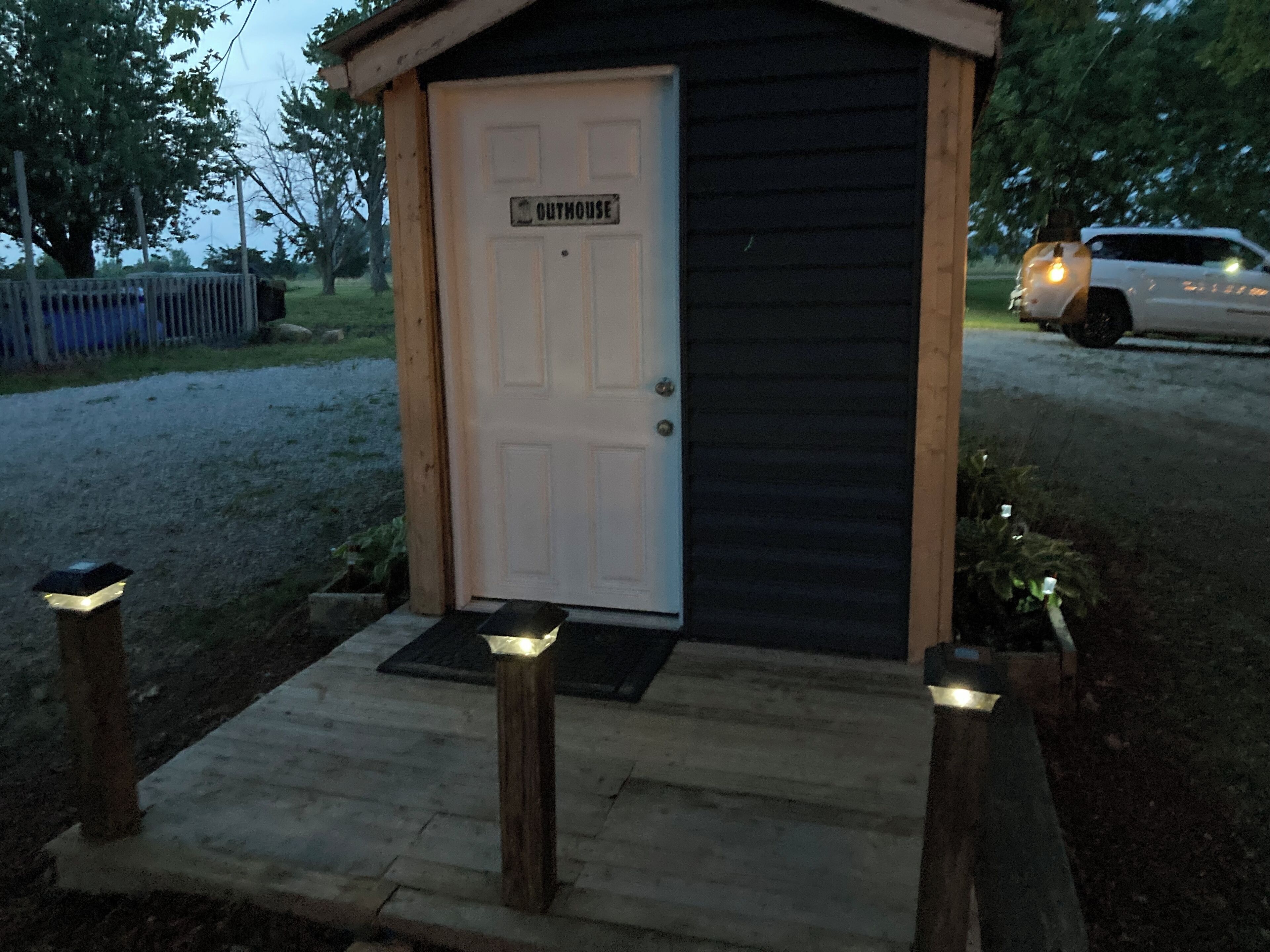 Een klein, gelijkvloers gebouw met een witte deur waarop 'OUTHOUSE' staat. Het heeft een donkere buitenkant en een houten veranda met drie lampen.