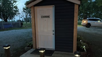Een klein, gelijkvloers gebouw met een witte deur waarop 'OUTHOUSE' staat. Het heeft een donkere buitenkant en een houten veranda met drie lampen.