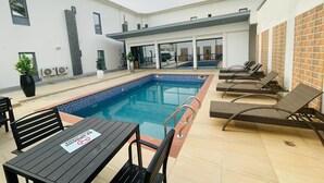 A modern outdoor pool area with lounge chairs and a table, surrounded by a white building with a brick accent wall.