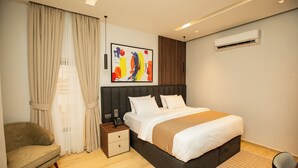 A modern hotel room with a large bed, a bedside table with a phone, a chair, and a large abstract painting on the wall.