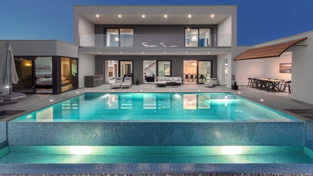 A modern two-story house with a large swimming pool, outdoor seating, and a dining area.