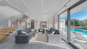 A modern living room with a staircase, grey sofas, a coffee table, and a view of a swimming pool.