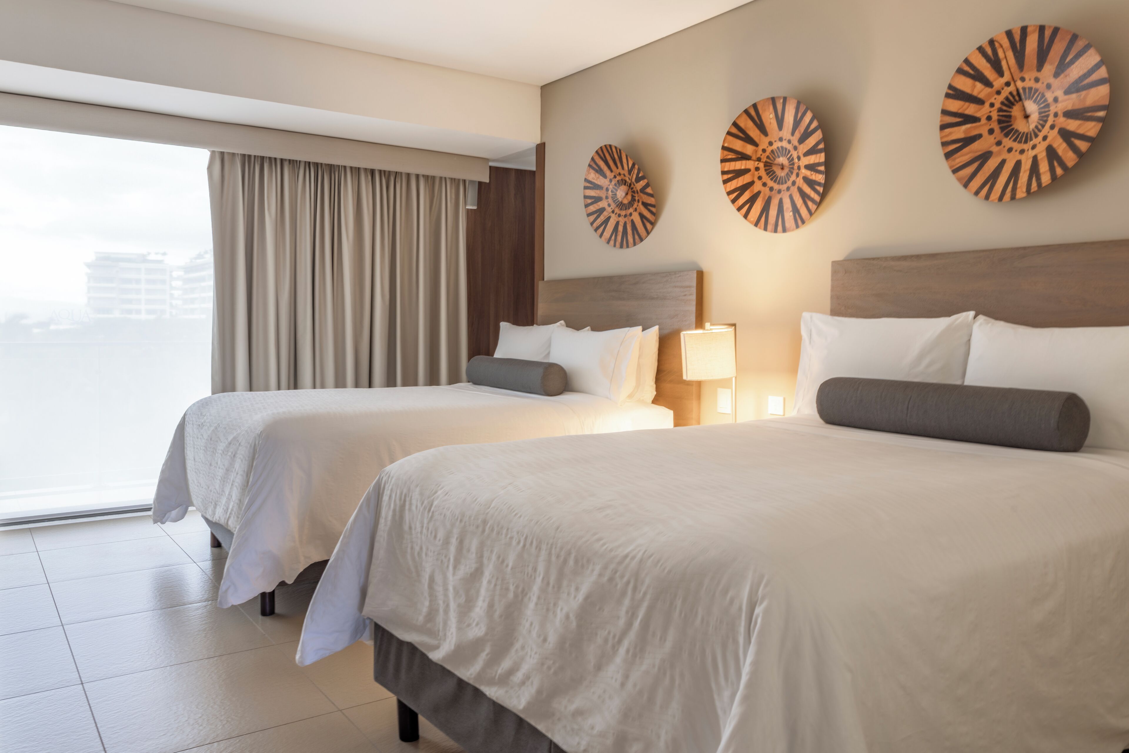 Una habitación de hotel con dos camas, un ventanal con cortinas y tres relojes en la pared.