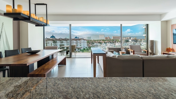 A modern living room with a large window, a wooden dining table, and a sofa. - Live Aqua Private Residences Vallarta (Puerto Vallarta)