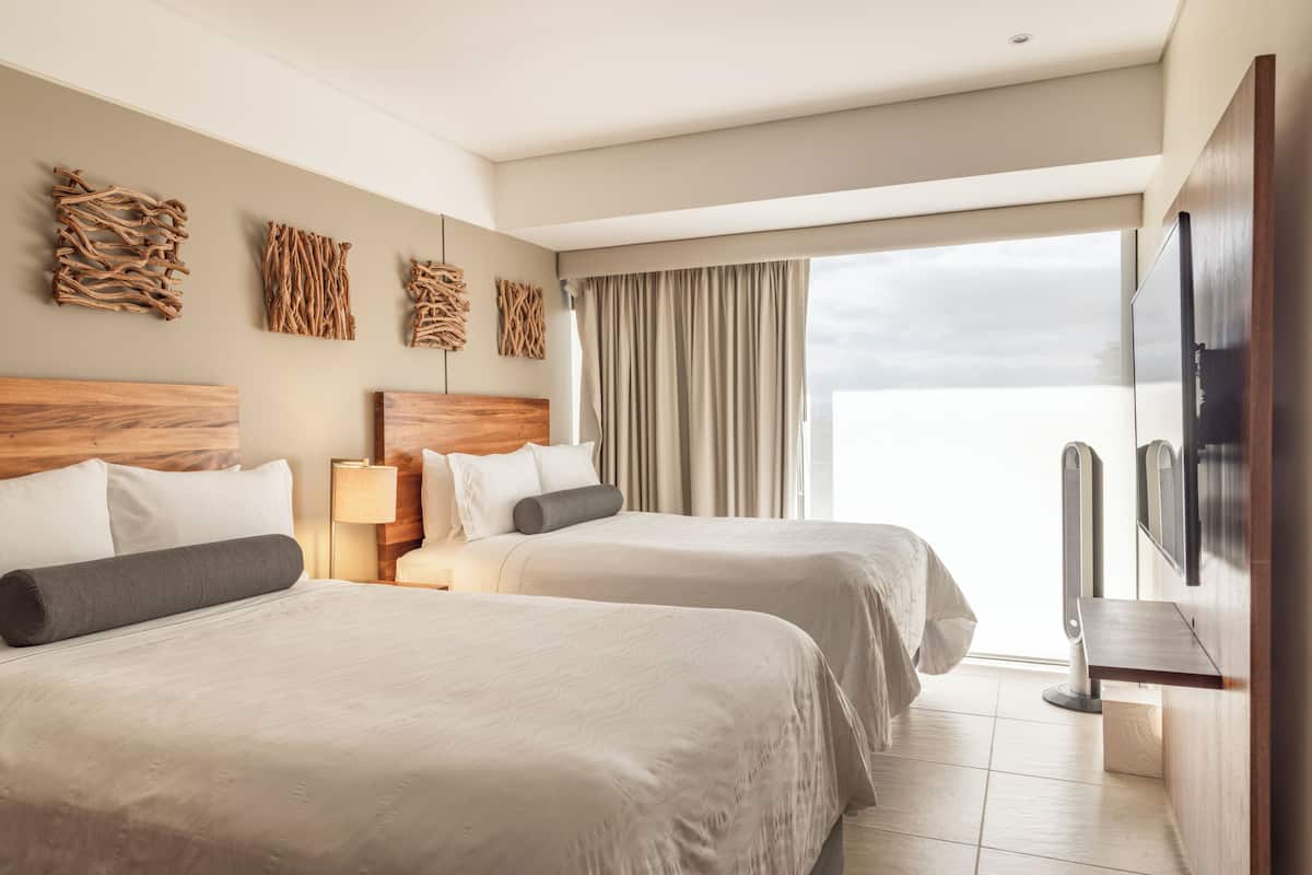 A hotel room with two beds, a TV, a desk, and a large window with curtains.