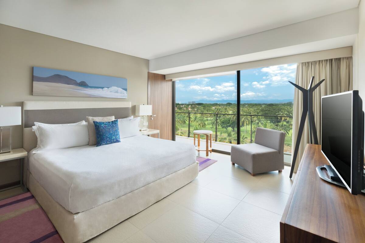 A modern hotel room with a large bed, a TV, a desk, and a balcony with a view of greenery.