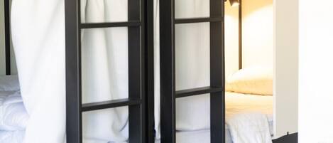 A bunk bed with white bedding and curtains, a bedside lamp, and a view of a bed with a pillow.