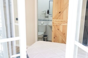 A bedroom with a bed, a wooden door, a small window, and a view of a bathroom with a toilet, sink, and mirror. - HOTEL NAUTICO (Vigo)