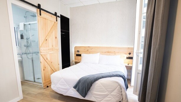 A bedroom with a wooden bed, a sliding door to a bathroom, and a view of a building outside. - HOTEL NAUTICO (Vigo)