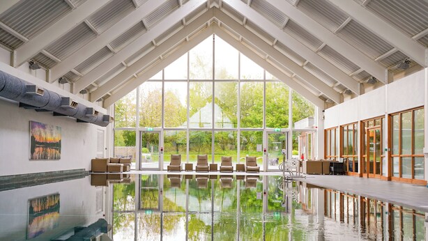 A large indoor swimming pool with clear glass walls, a high ceiling with skylights, and a view of greenery outside. - Bauhinia House: Spa Access and Lakeside Home, Lower Mill Estate (Somerford Keynes)