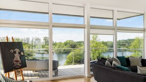 A spacious balcony with a view of a lake, a painting on easel, and a dark-colored sofa with cushions. - Bauhinia House: Spa Access and Lakeside Home, Lower Mill Estate (Somerford Keynes)