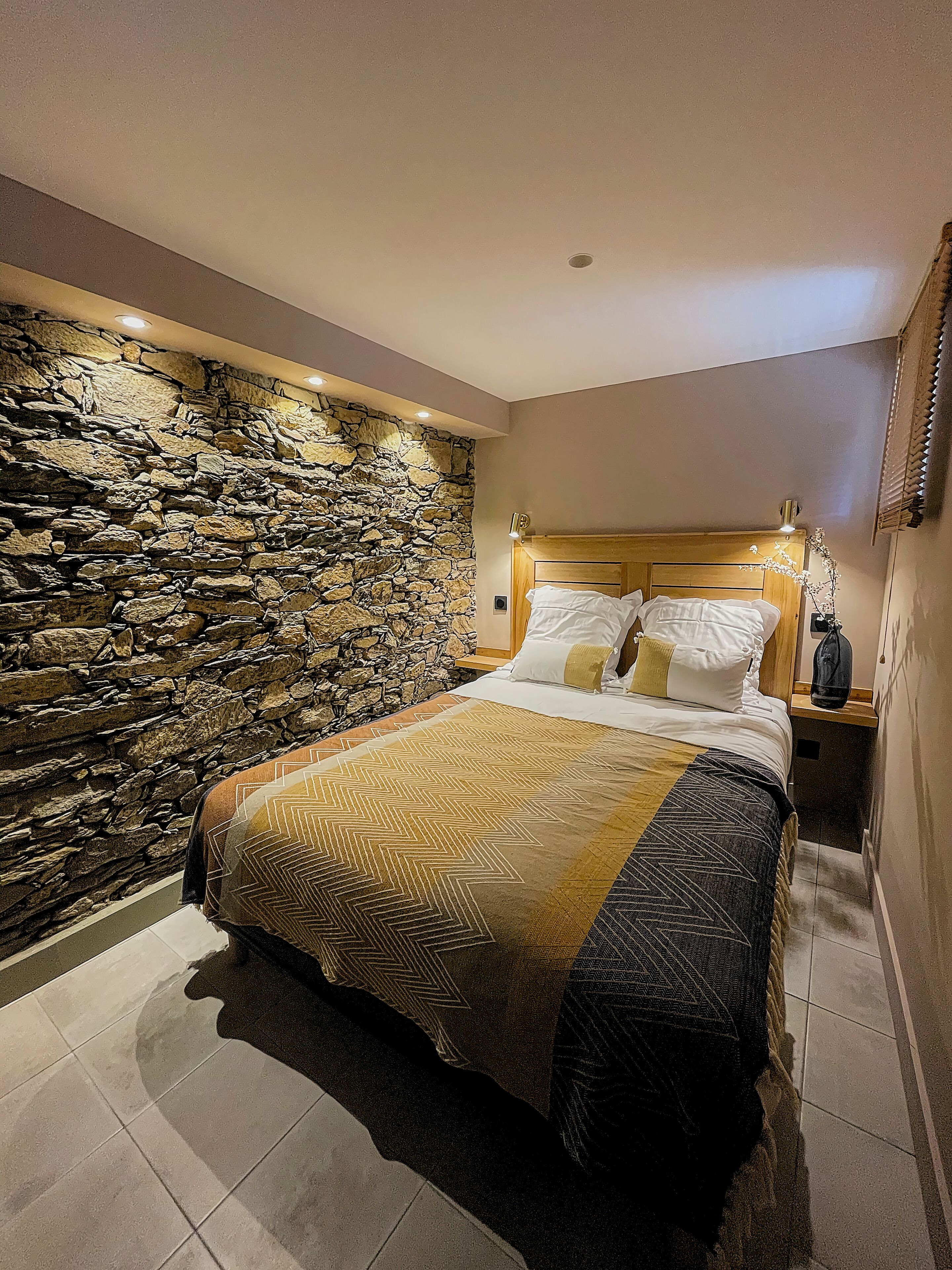 A bedroom with a stone wall, a bed with patterned bedding, and a nightstand with a lamp.