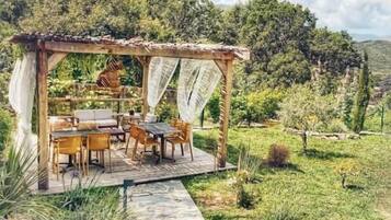 Un sendero de piedra conduce a una pérgola de madera con una mesa y sillas, rodeado de exuberante vegetación y colinas al fondo.