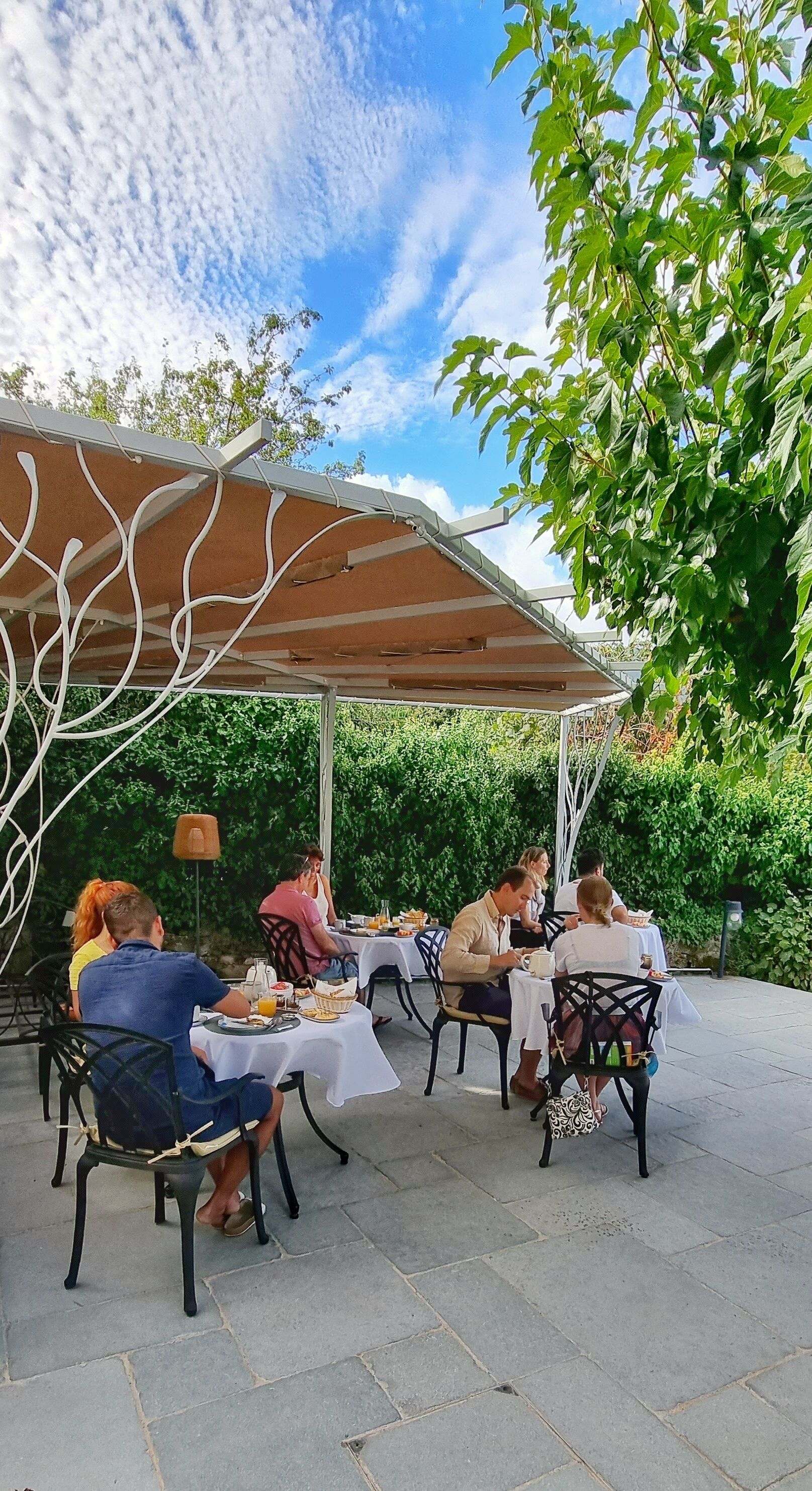 A patio area with tables and chairs under a pergola, surrounded by greenery.