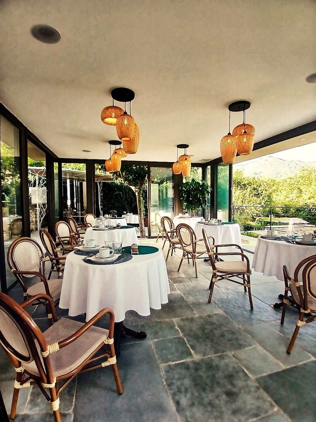 Un espace de restauration en plein air avec des tables rondes et des chaises en osier, entouré de grandes fenêtres offrant une vue sur la verdure.