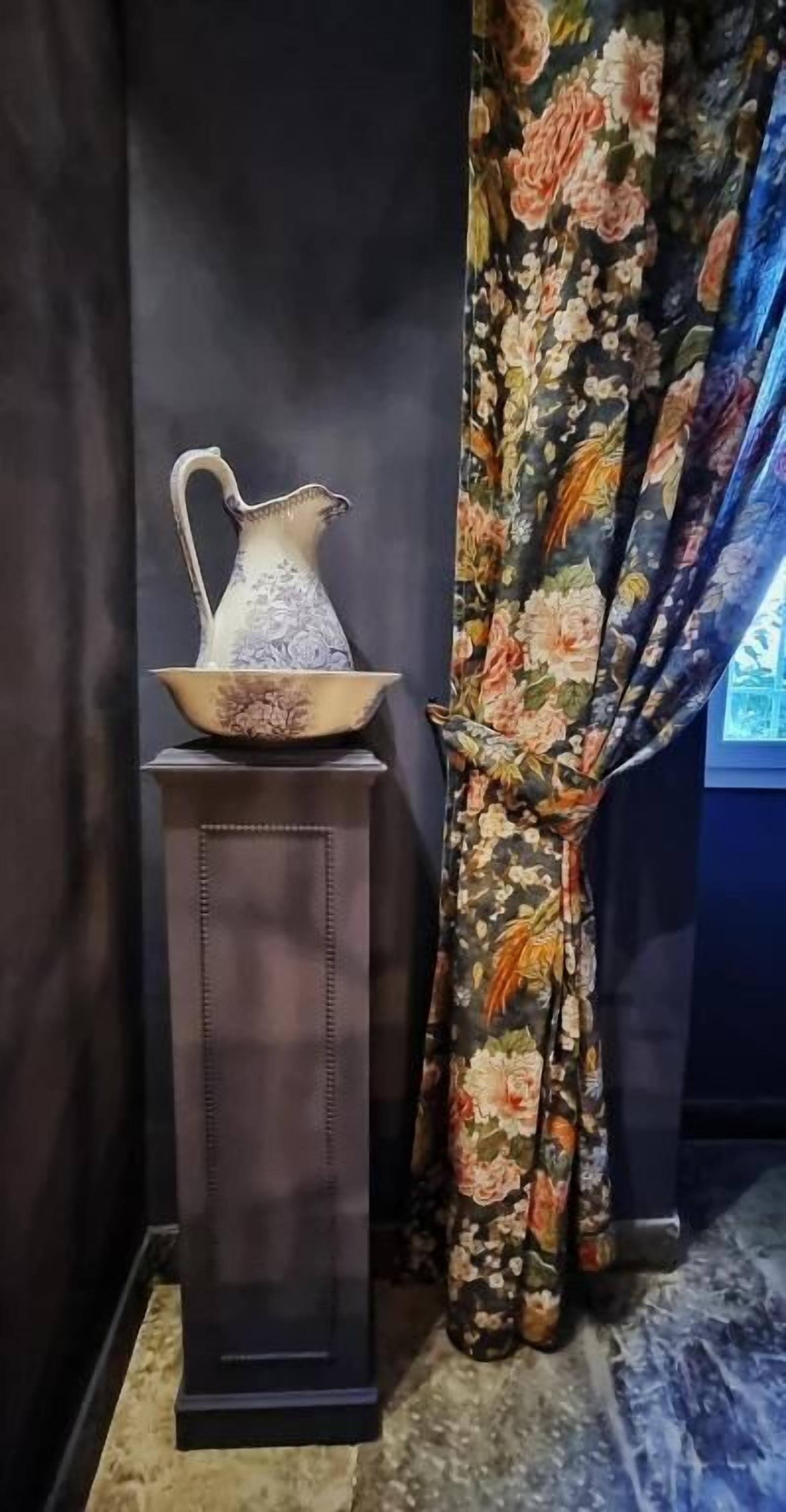 Una mesa oscura con una jarrón y un cuenco encima, junto a una ventana con cortinas florales.