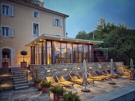 Una villa moderna con un porche acristalado, escalones de piedra y mobiliario exterior con sombrillas.