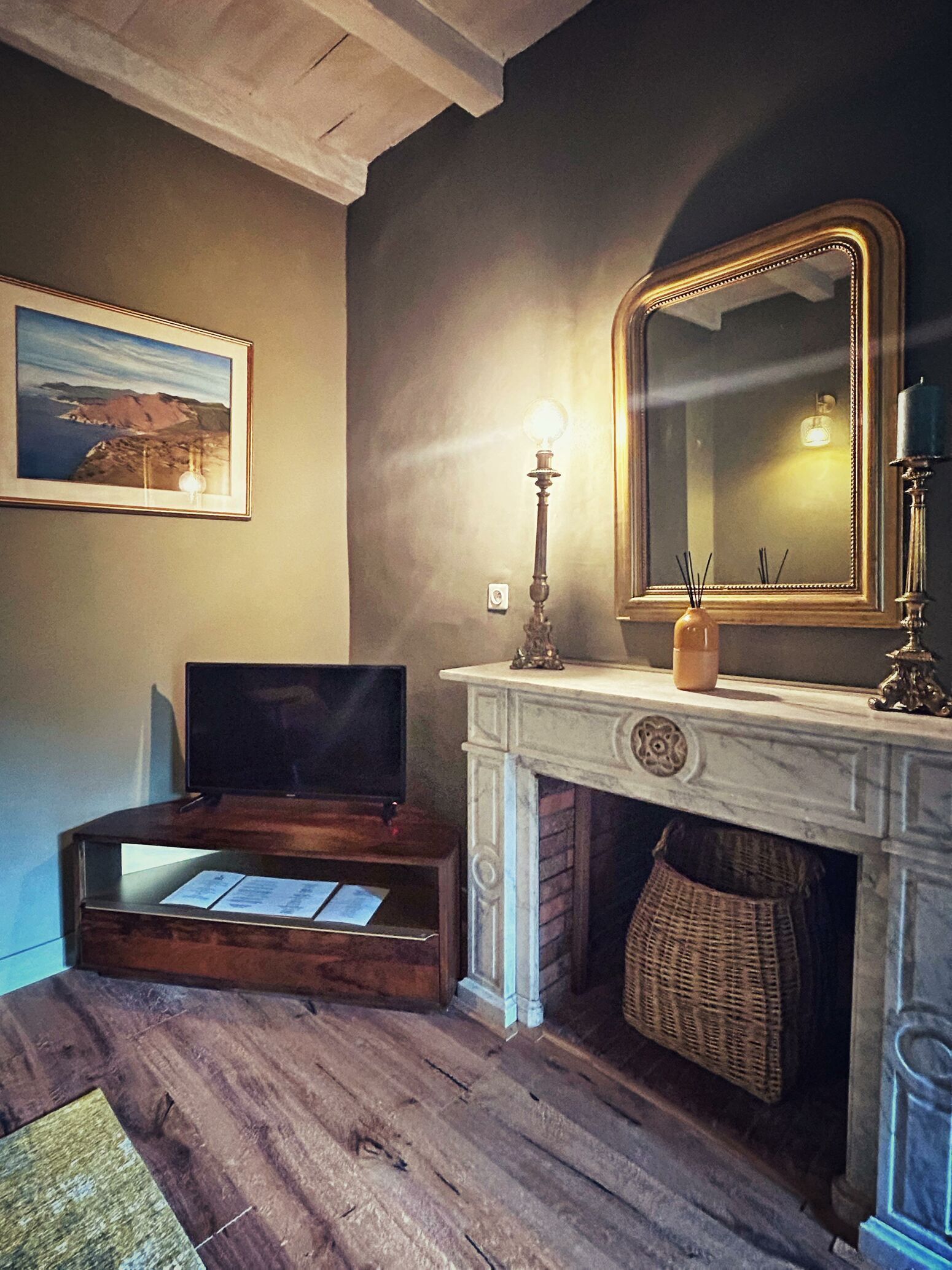 A room with a fireplace, a framed mirror, a television on a wooden stand, and a framed picture on the wall.