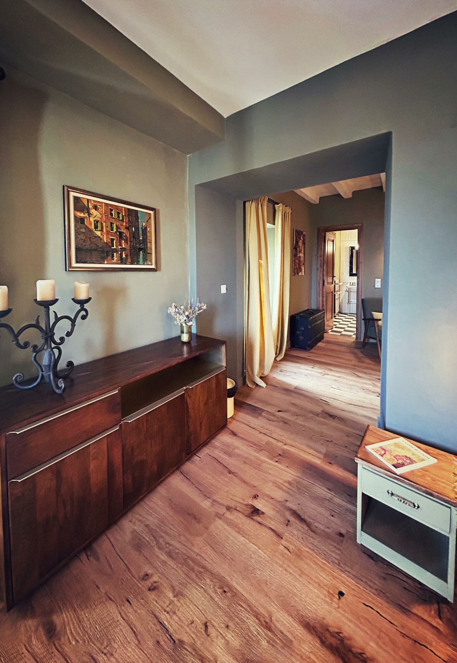 Un couloir doté d’un plancher en bois, une console en bois sombre surmontée d’un chandelier, un tableau encadré accroché au mur et une petite table avec un livre.