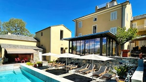 A swimming pool with lounge chairs and umbrellas, a stone wall, and a large building with multiple windows. - À Villa di Rutali (Rutali)