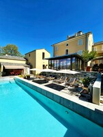 Una piscina con sillas de descanso y sombrillas, un muro de piedra y un edificio grande con numerosas ventanas.