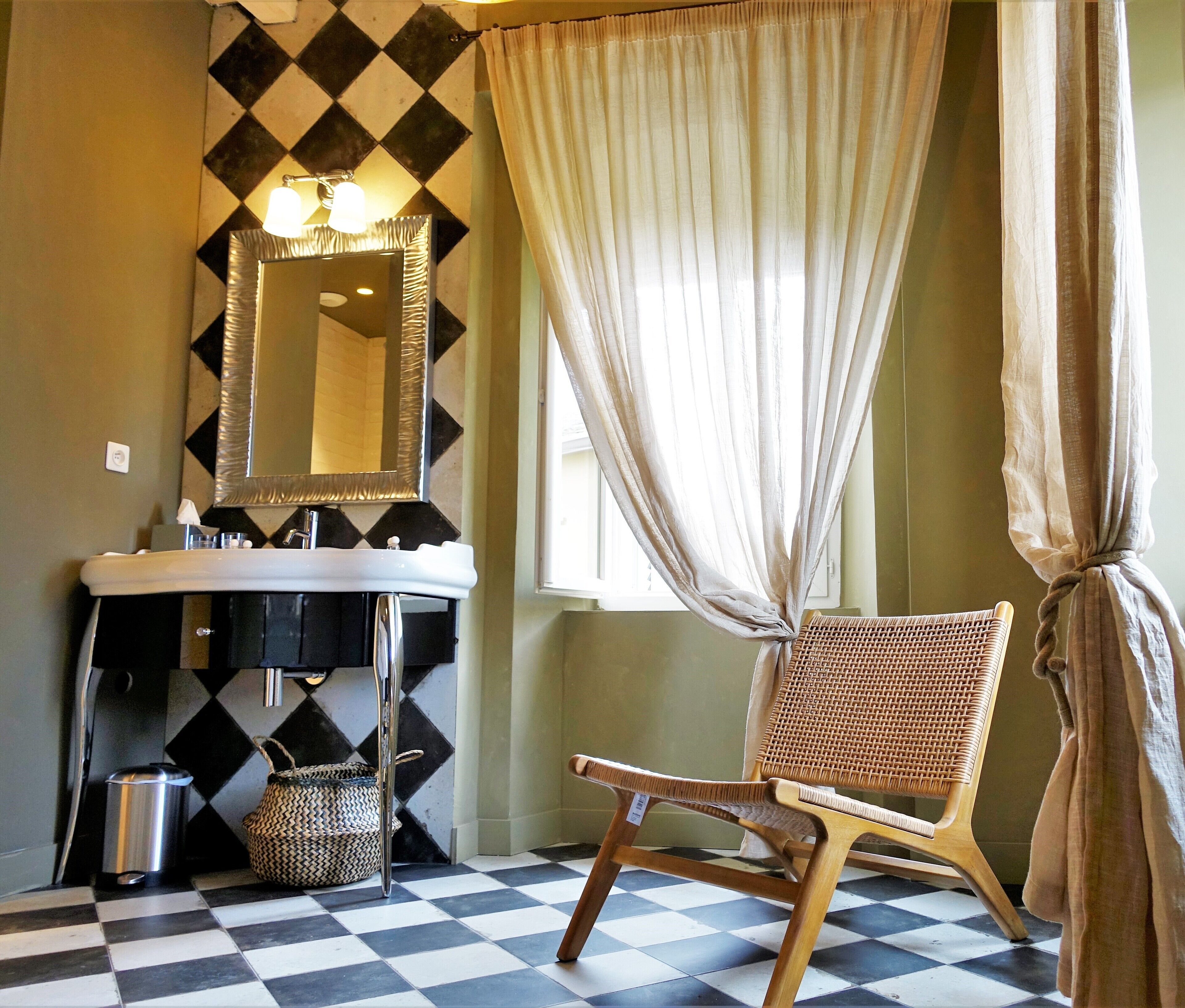 A bathroom with a checkered floor, a sink, a mirror, a wicker chair, and a window with curtains.