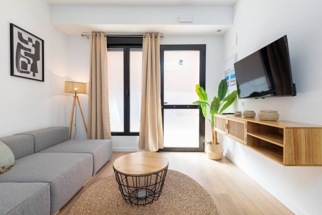 Ein modernes Wohnzimmer mit grauem Sofa, einem runden Couchtisch, einem an der Wand montierten Fernseher und einem großen Fenster mit Vorhängen.