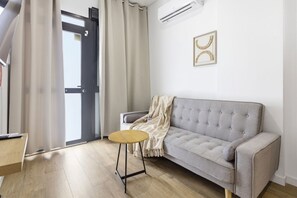 Ein modernes Wohnzimmer mit einem grauen Sofa, einem runden Holztisch, einer beigefarbenen Decke und einem gerahmten Bild an der Wand.