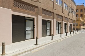 A row of closed storefronts with metal shutters, a sidewalk, and bollards along a street.