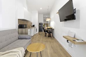 Eine moderne Küche mit Einbauofen, ein Essbereich mit Holztisch und Stühlen, eine Couch und ein an der Wand befestigter Fernseher.