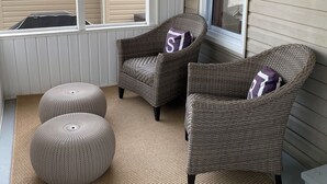 Een overdekt terras met rieten stoelen en poefs, een witte deur en een raam.