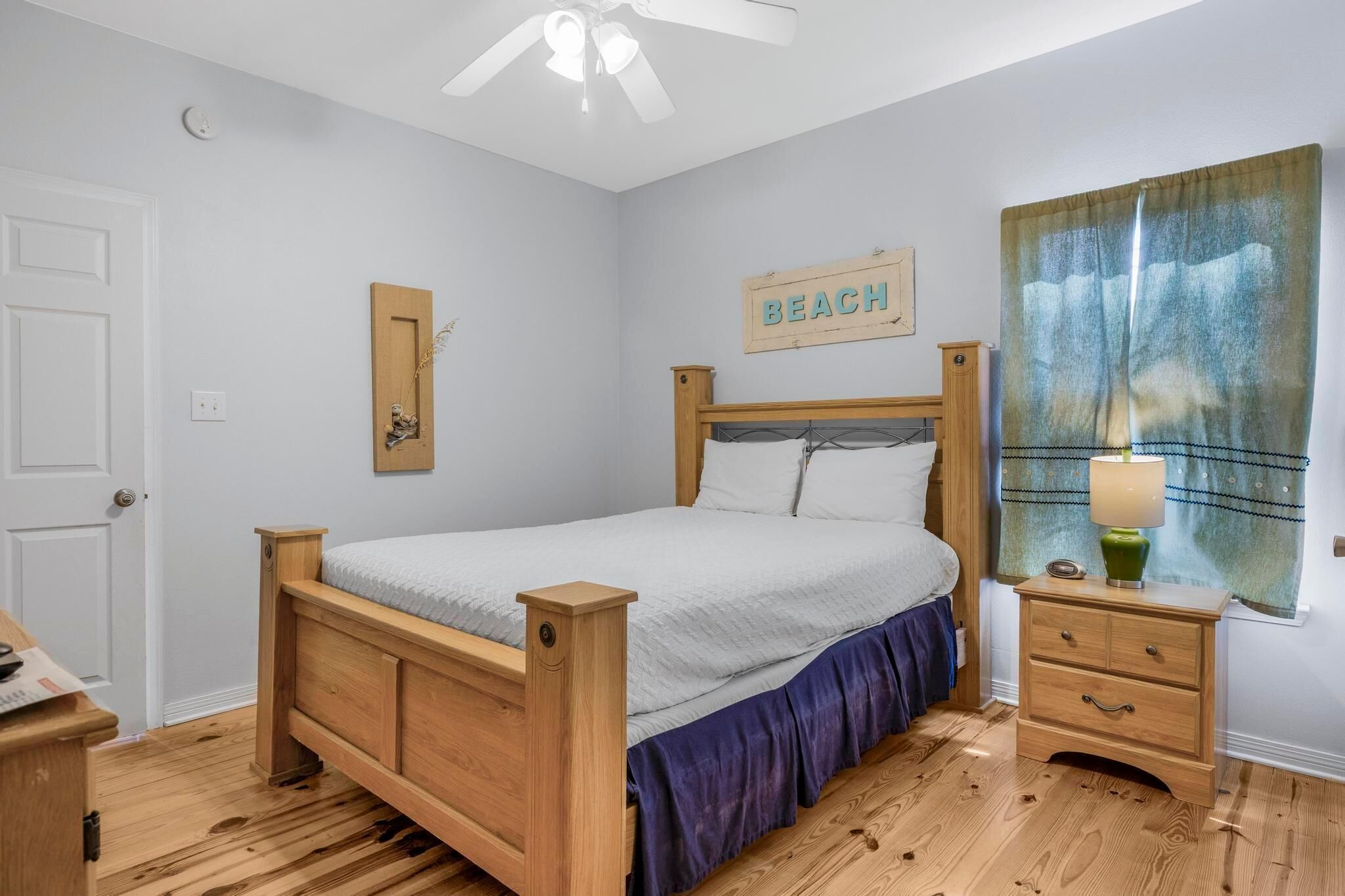 A bedroom with a wooden bed, a nightstand, a lamp, a window with a beach-themed curtain, and a 'BEACH' sign on the wall.