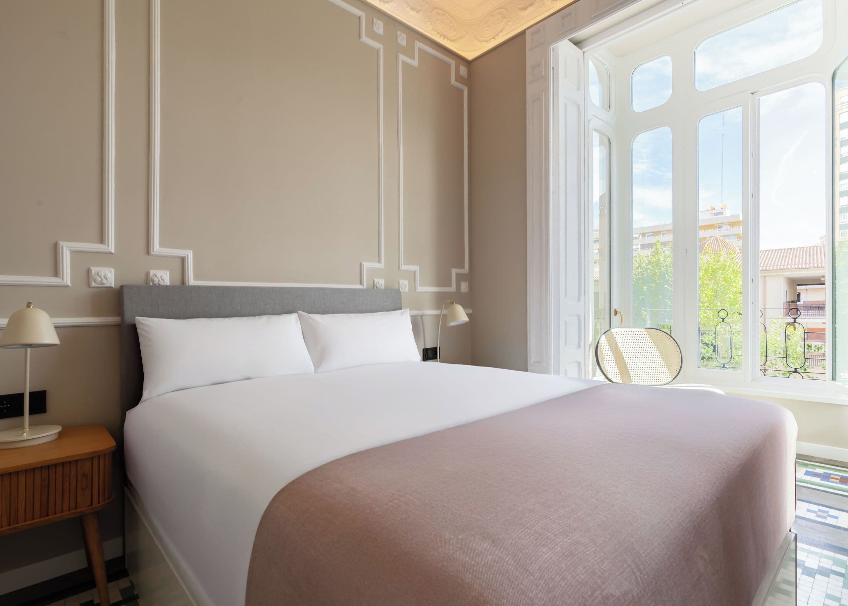 A neatly made bed with a grey headboard and white bedding, a bedside table with a lamp, and a window with a view of buildings and greenery.