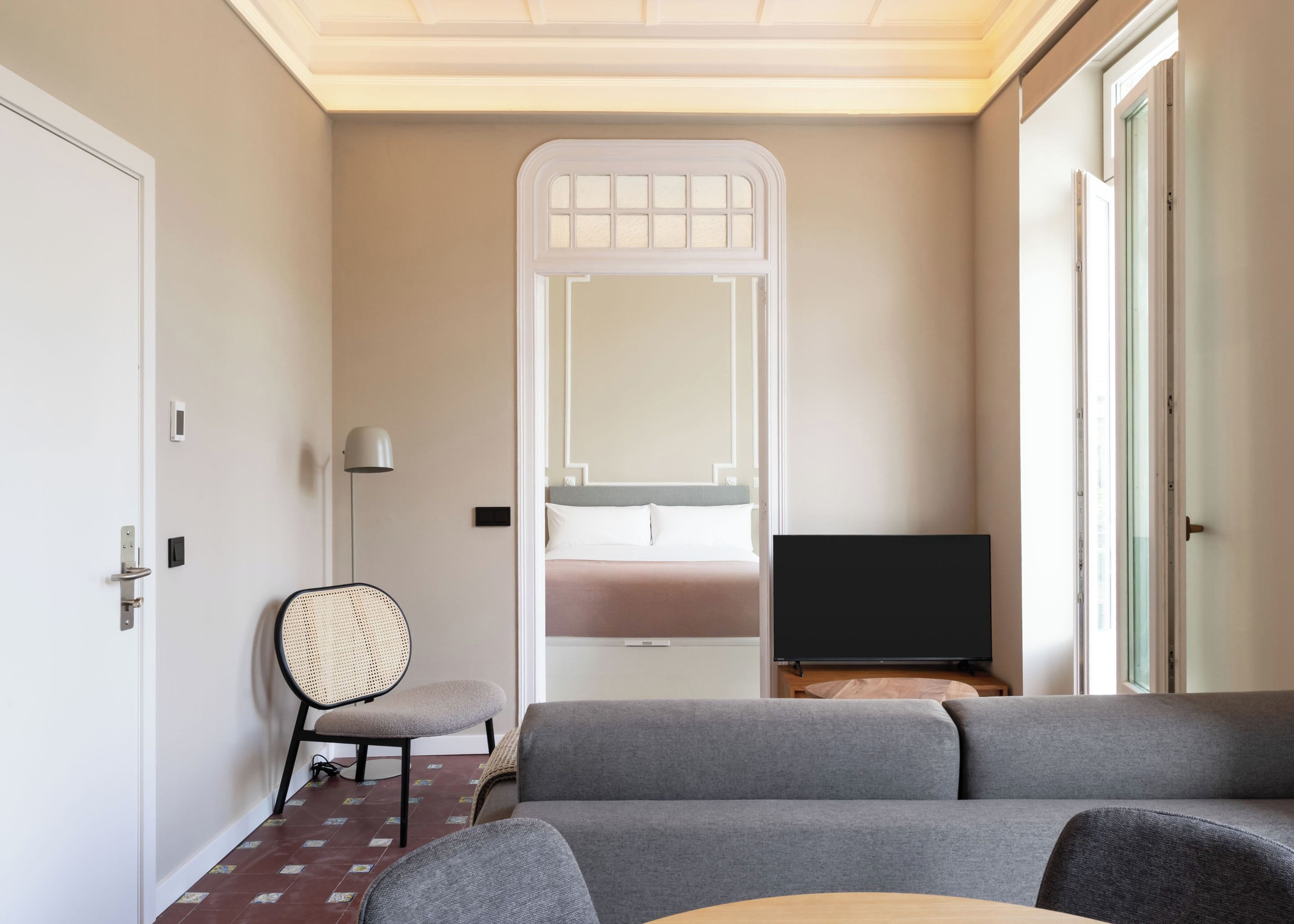 A modern hotel room with a grey sofa, a small round table, a rattan chair, a television, and a large mirror on the wall.