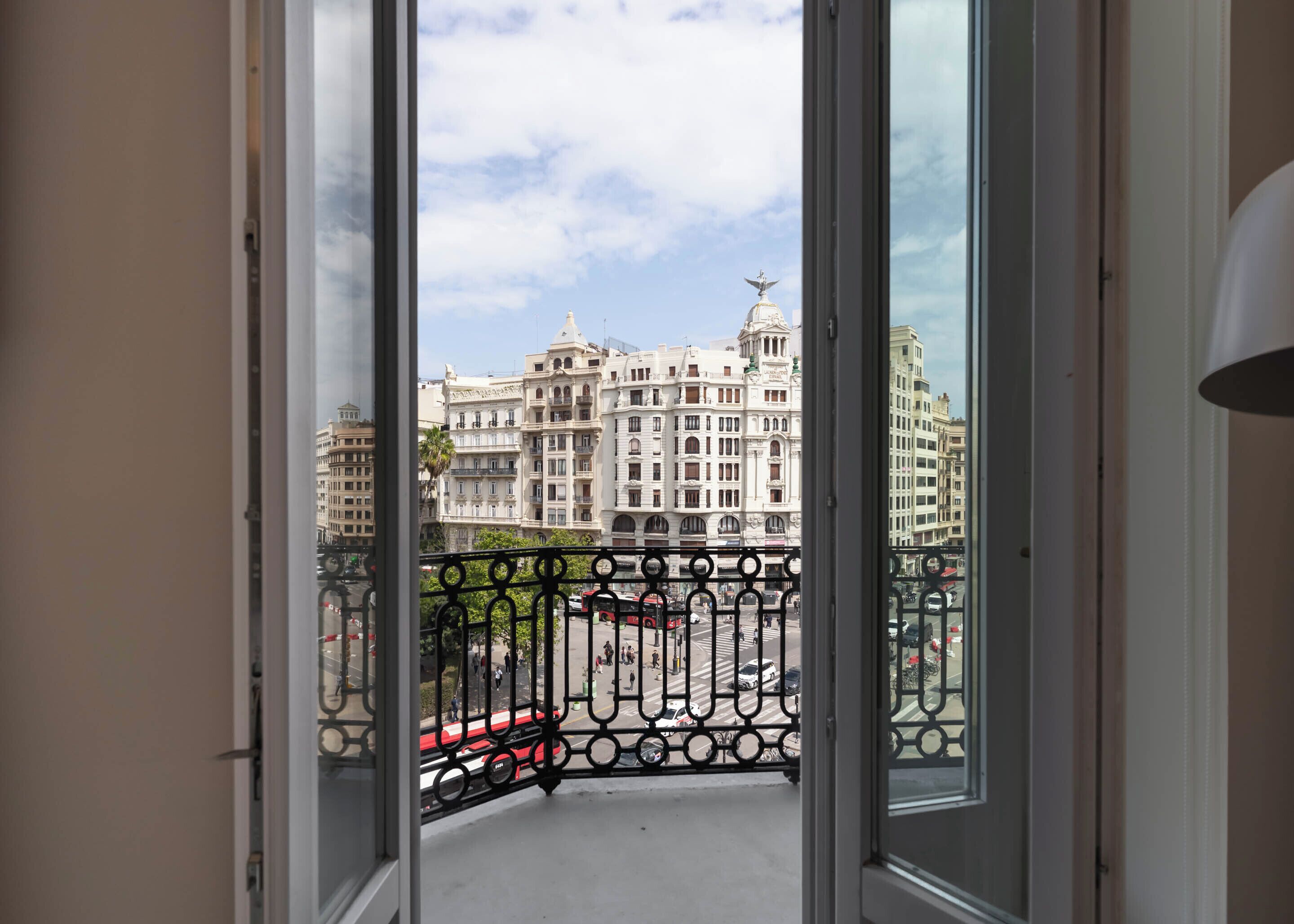 Vista desde un balcón con barandilla de hierro forjado, que da a una calle con coches y un edificio grande y ornamentado de arquitectura clásica.