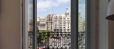 Vista desde un balcón con barandilla de hierro forjado, que da a una calle con coches y un edificio grande y ornamentado de arquitectura clásica.