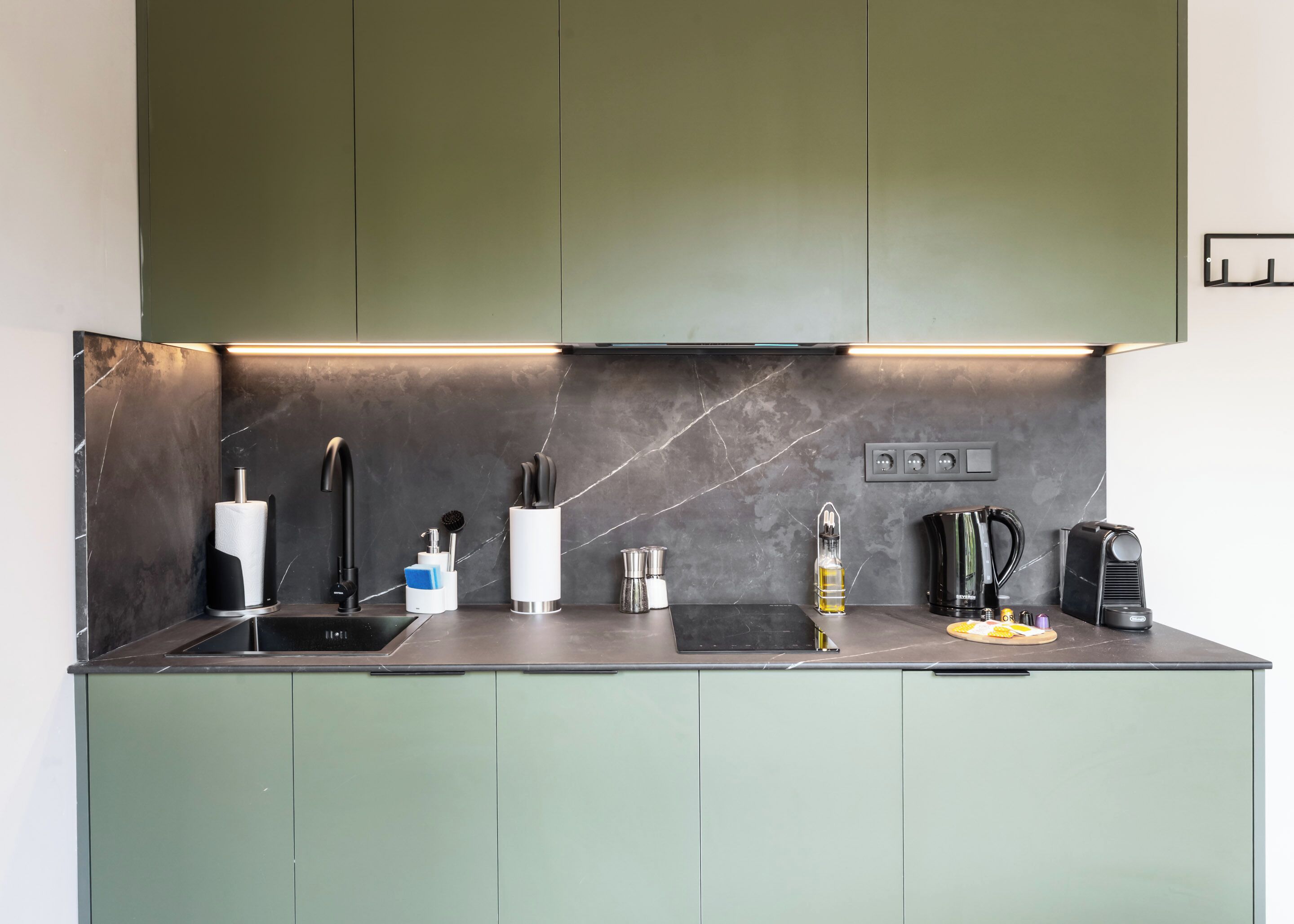 A modern kitchen with a dark countertop, green cabinets, and a stainless steel sink.