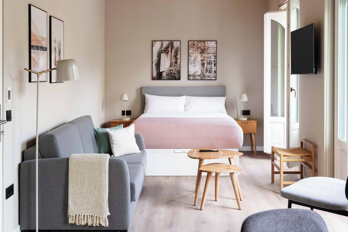 Een moderne slaapkamer met een bed, een grijze bank, een klein houten tafeltje en een aan de muur bevestigde televisie.