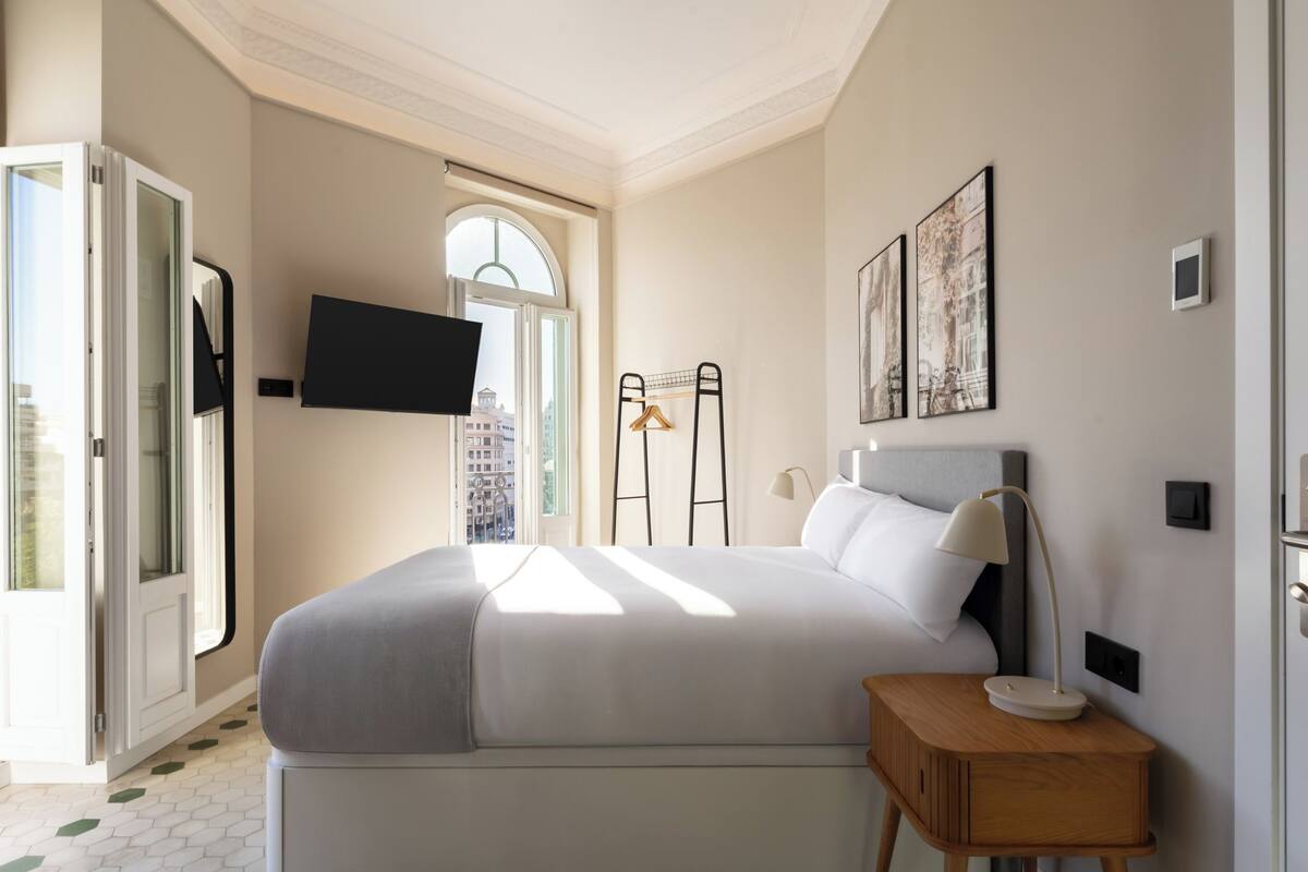 Een moderne slaapkamer met een groot bed, een nachtkastje, een televisie aan de muur en een raam met uitzicht op de stad.