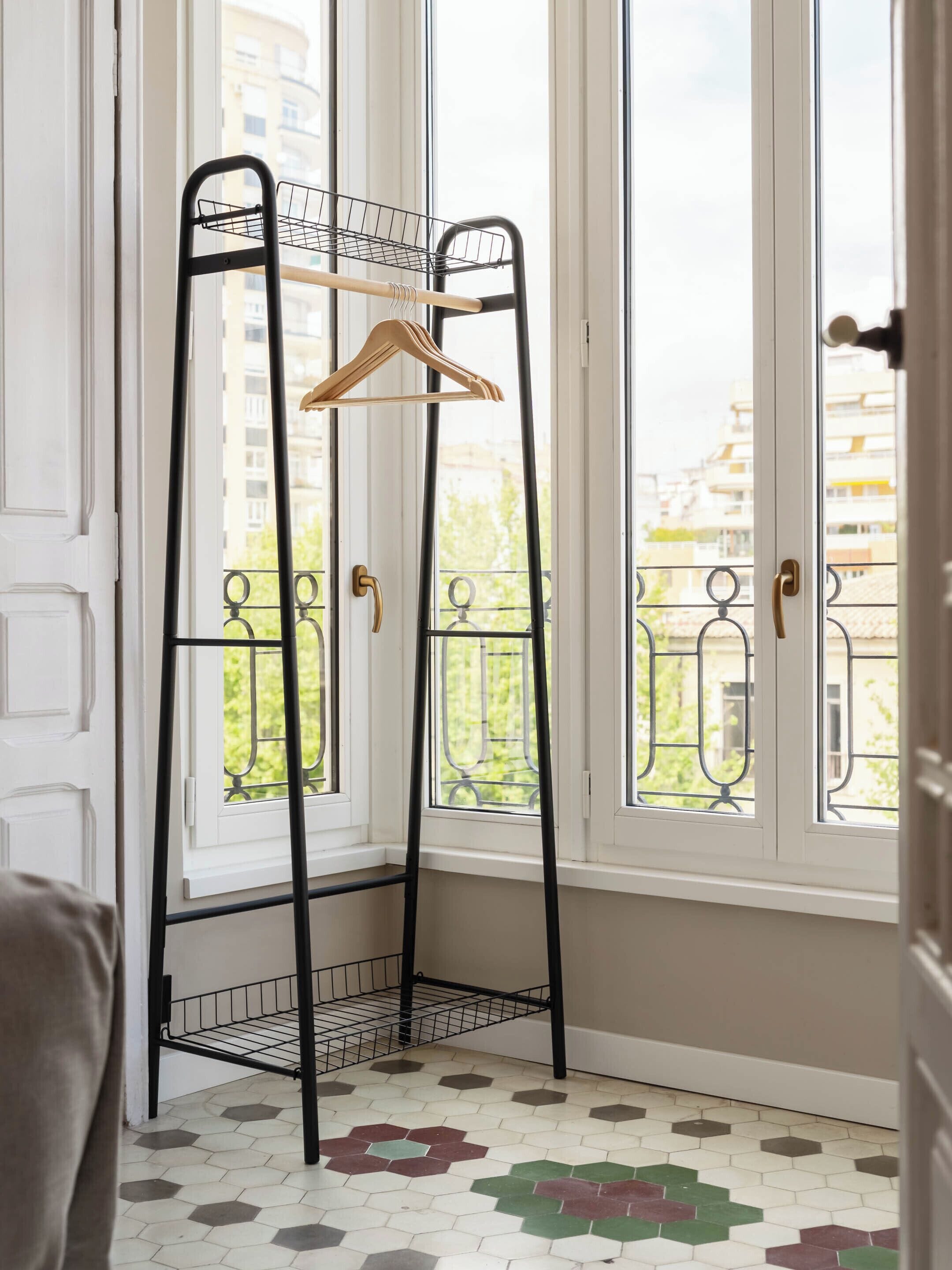 Una habitación de esquina con ventana, una estantería metálica con una percha de madera y suelo de baldosas.