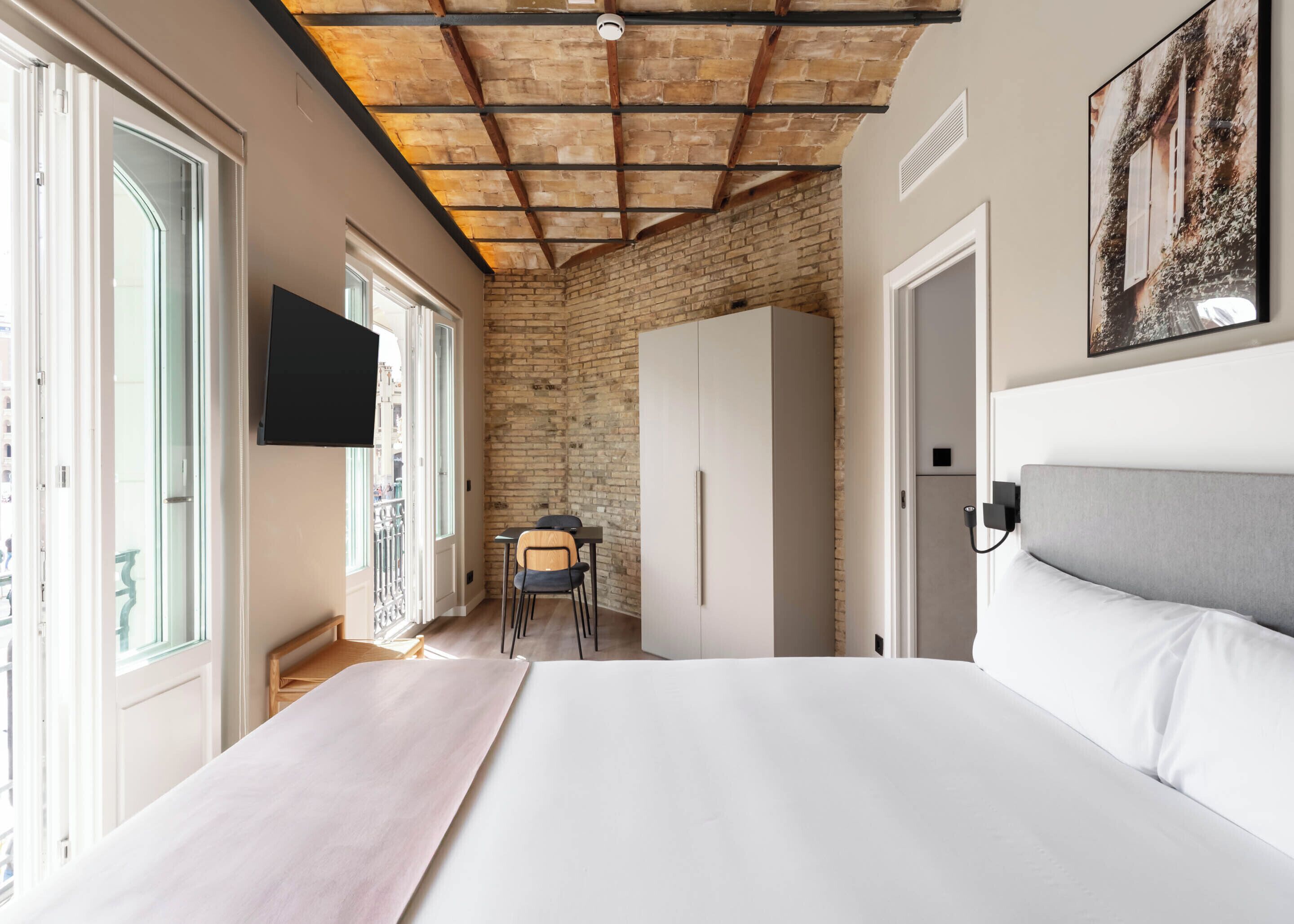 A bedroom with a large bed, a desk, and a chair, a TV mounted on the wall, and a brick wall.